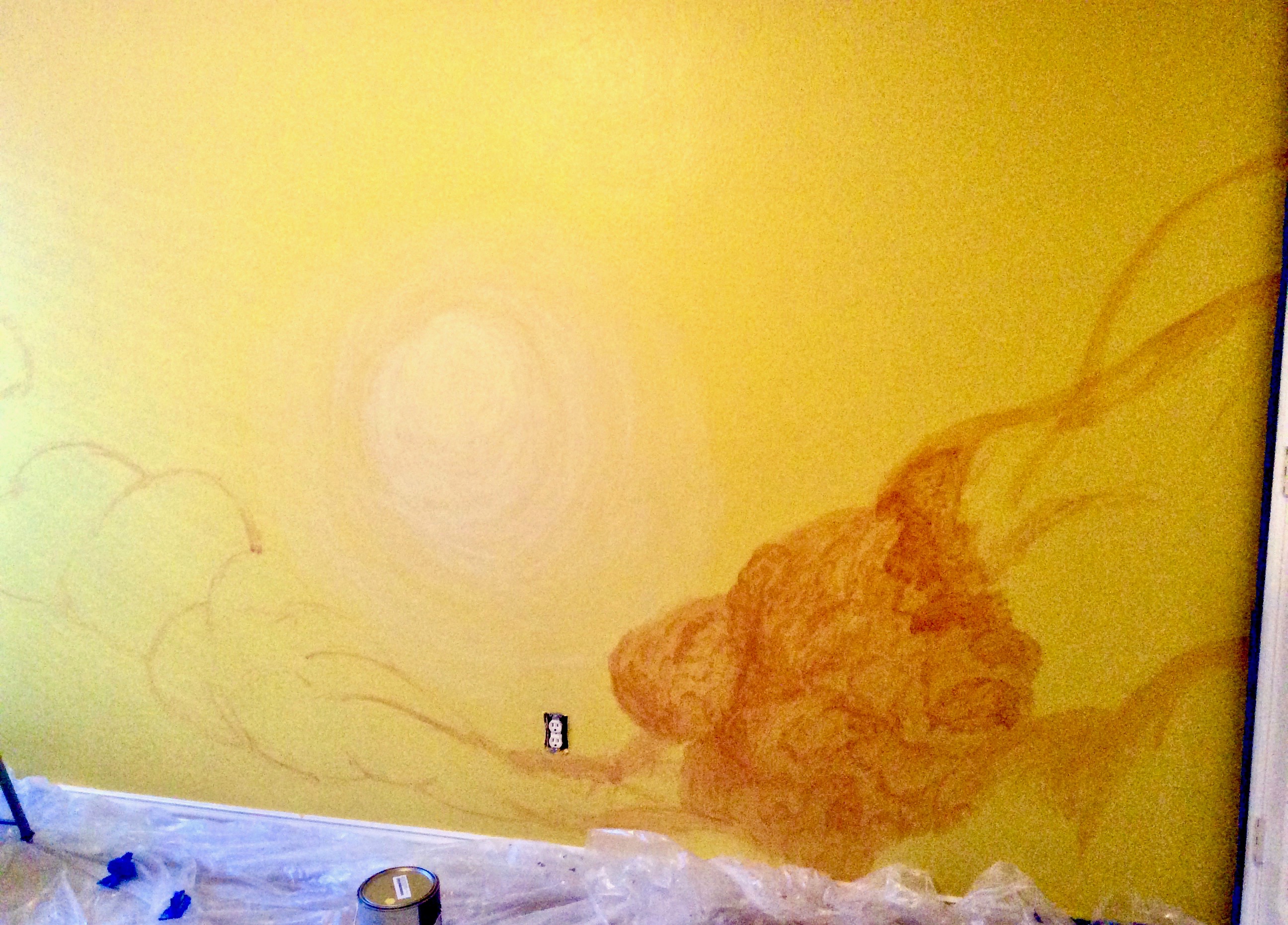 A pale yellow circular sun and the beginning of orange shaded clouds on a yellow wall 