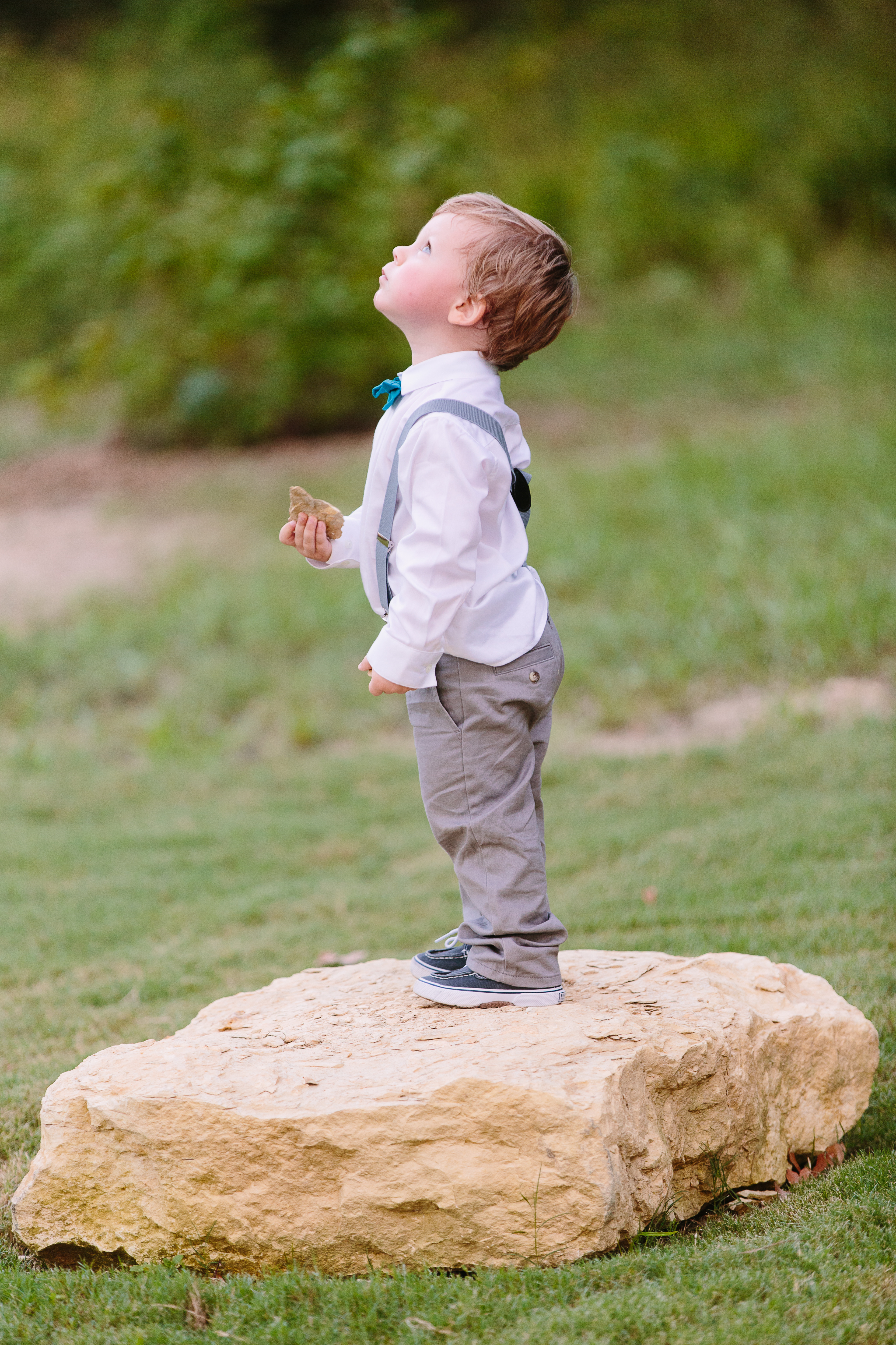 A toddler in dress clothes, a bowtie, and suspenders stands on a rock 