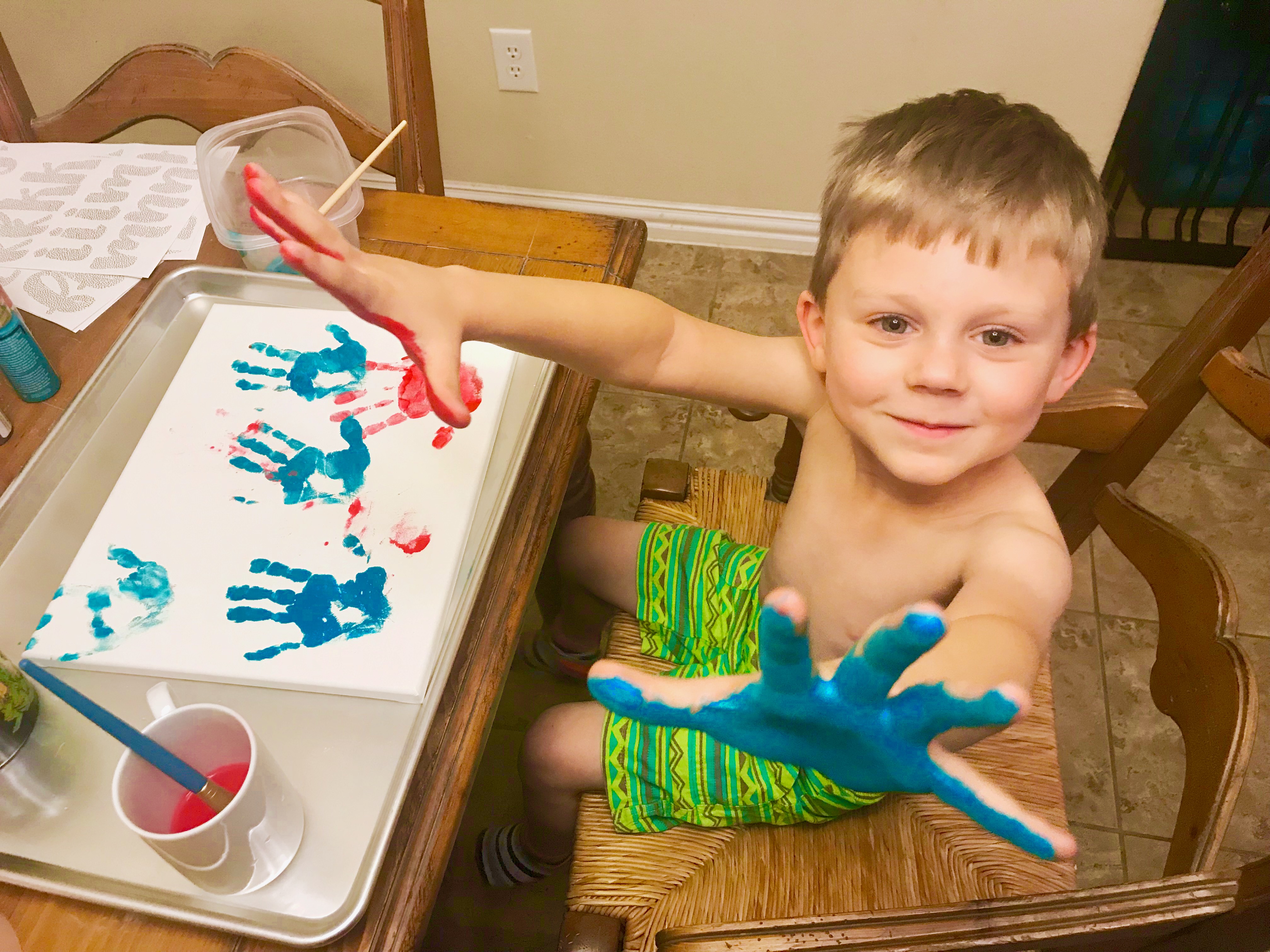 A child with brightly painted hands sitting in front of a canvas.