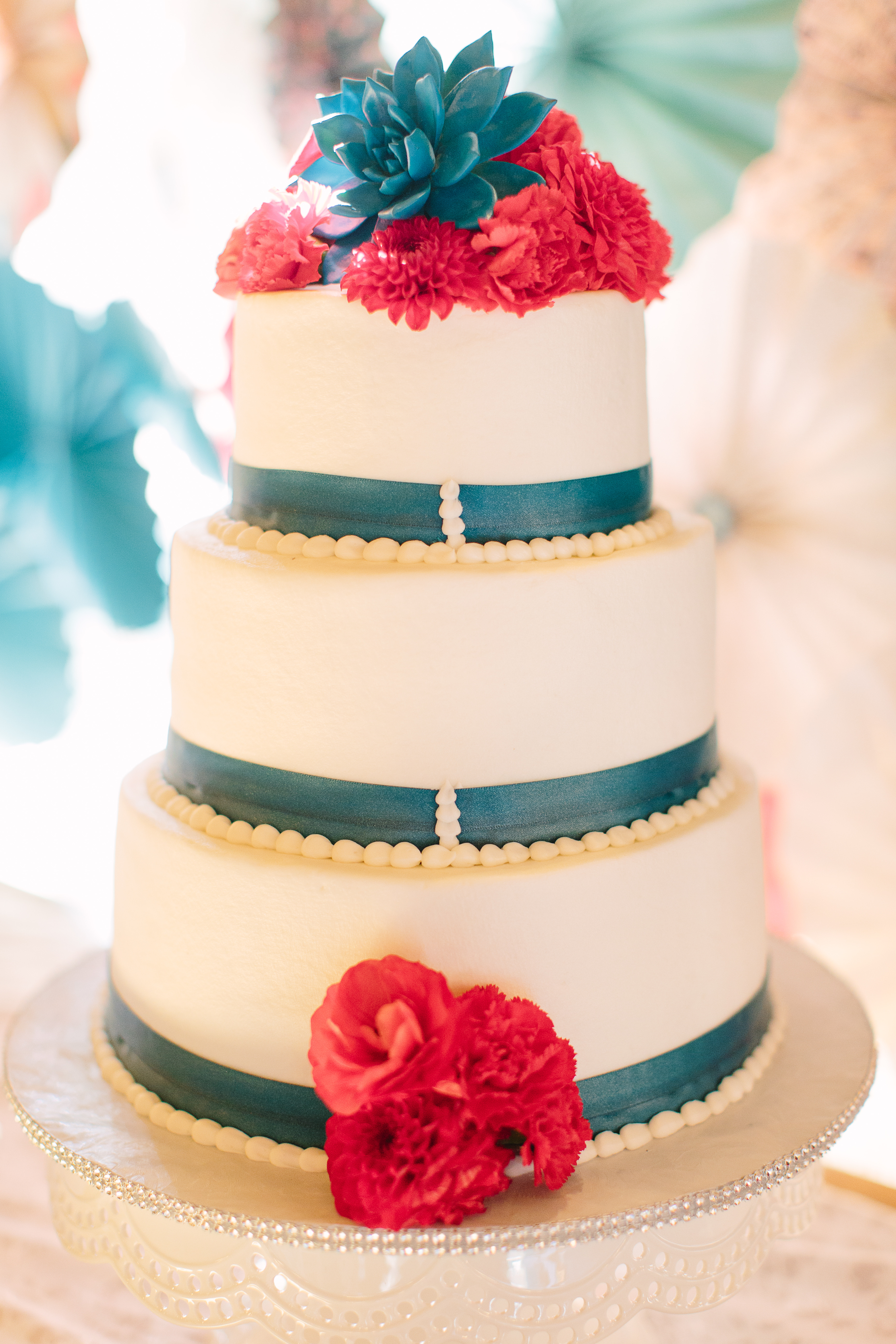 A 3-layer white buttercream wedding cake with teal ribbon, red flowers, and a teal succulent