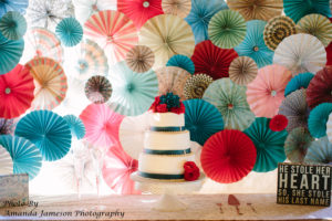 A wedding cake in front of a colorful paper pinwheel backdrop 