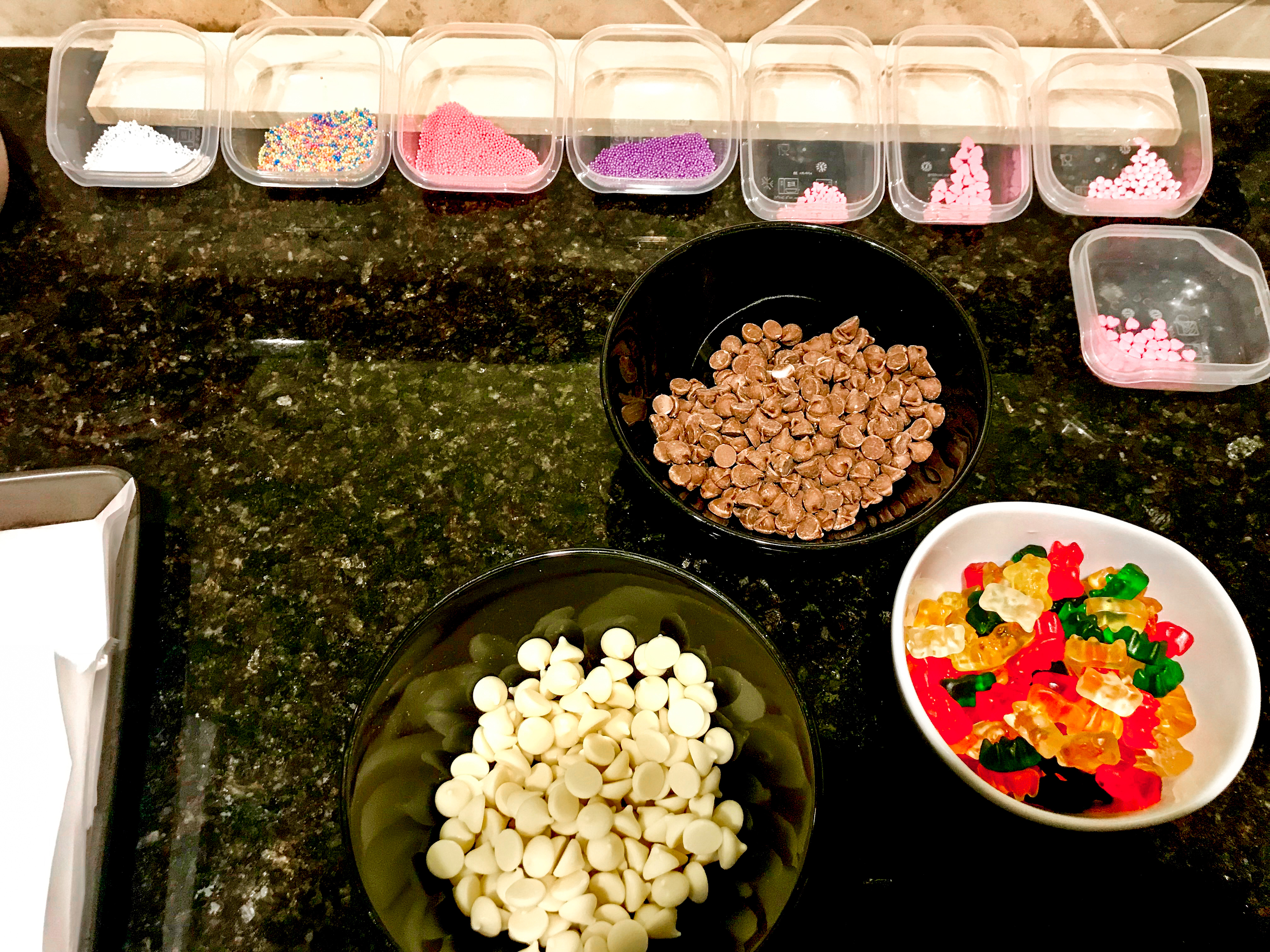 Bowls of chocolate chips, white chocolate chips, gummy bears, and sprinkles on a countertop. 