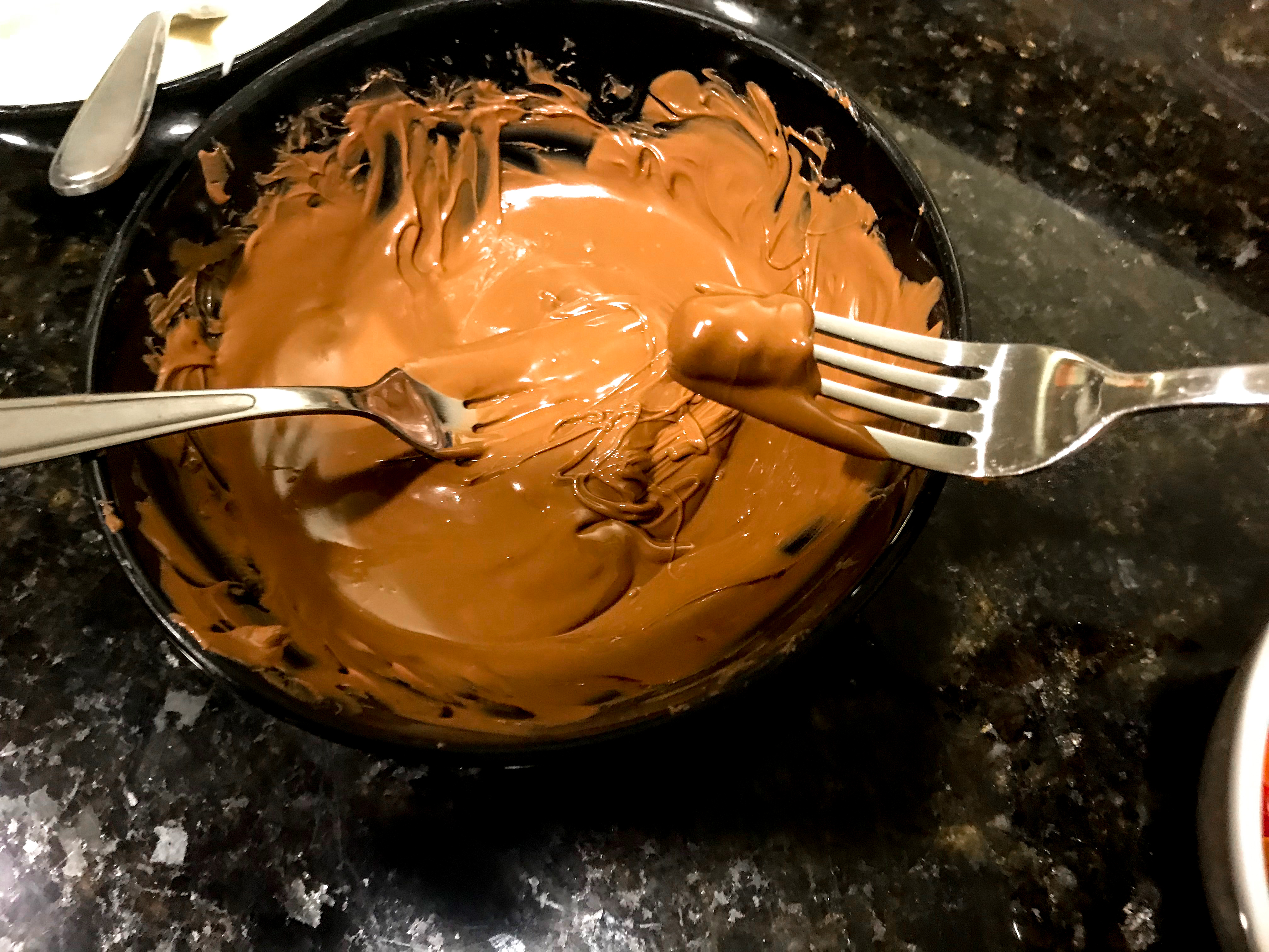 A bowl of melted milk chocolate and a recently dipped chocolate covered gummy bear on a metal fork. 