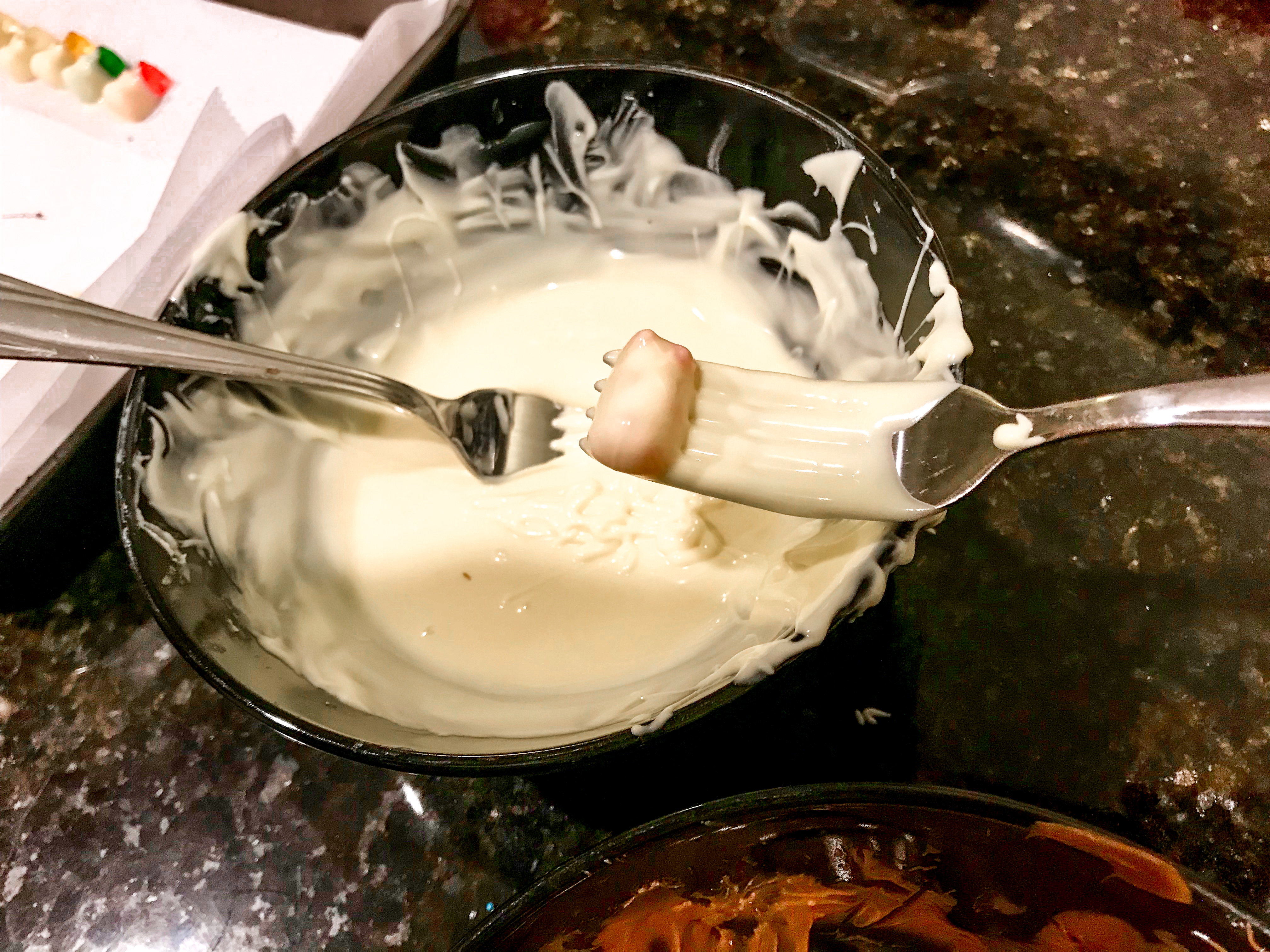 A bowl of melted white chocolate and a fresh white chocolate dipped gummy bear on a fork. 