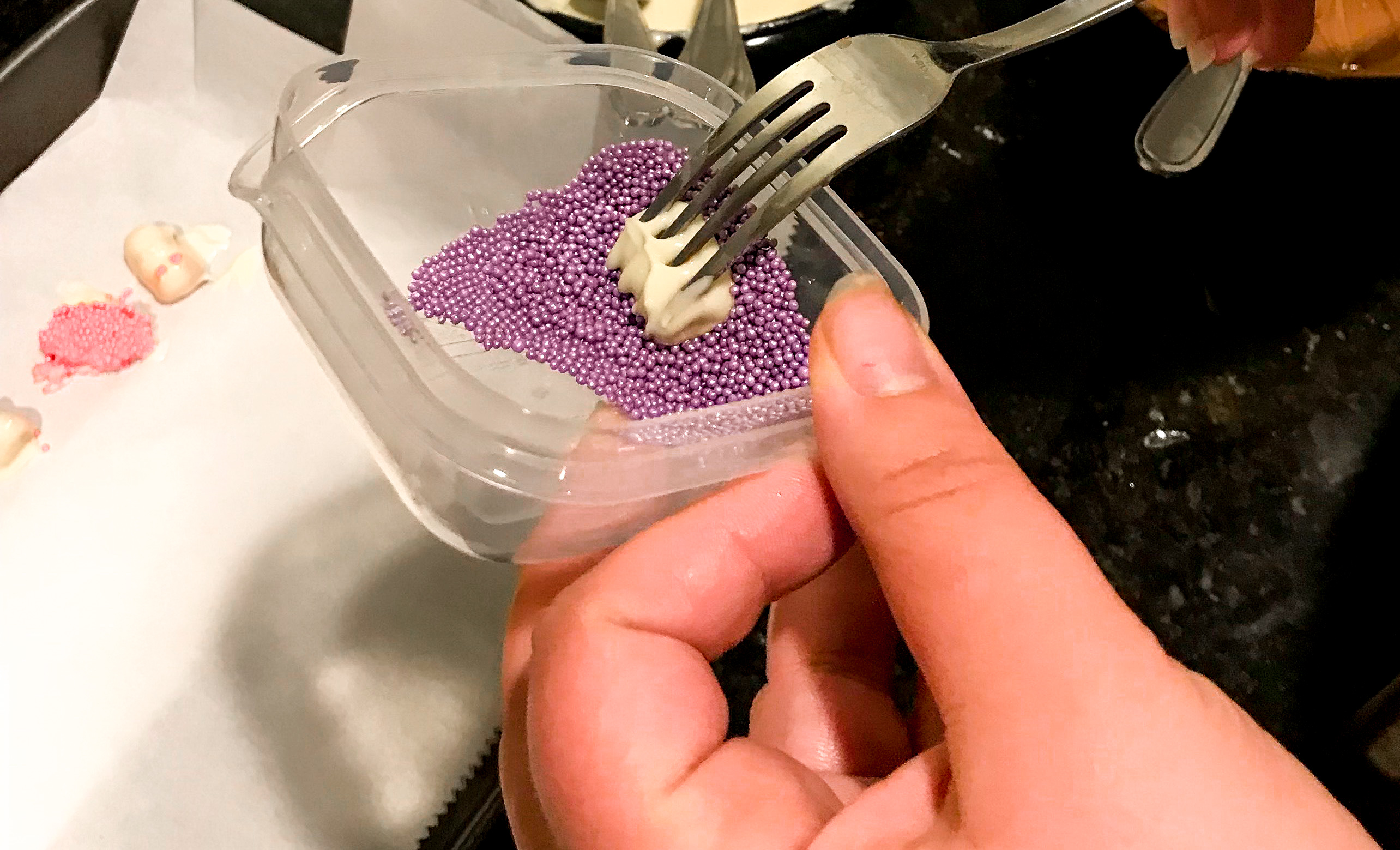 A white chocolate gummy bear on a fork being dipped in purple sprinkles