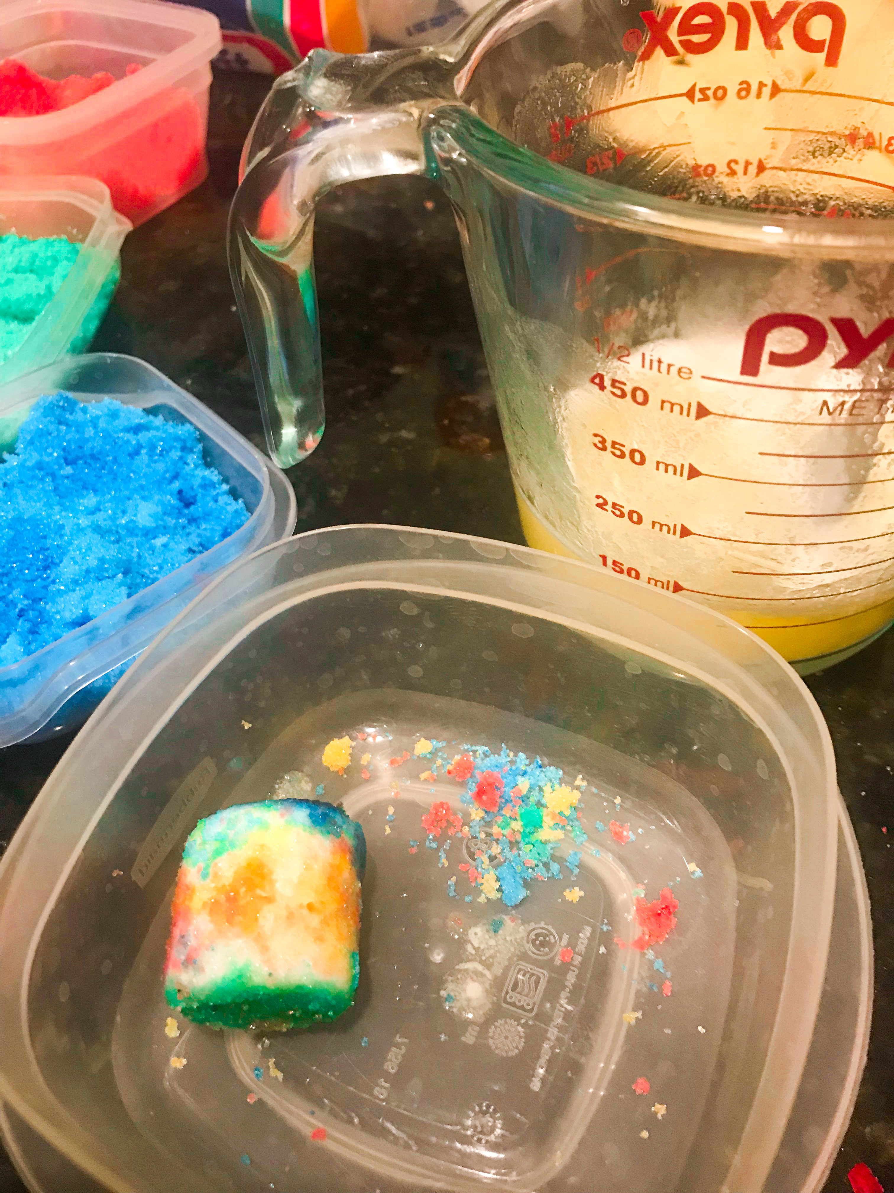 A marshmallow dipped in brightly colored sugar with melted butter and colored sugar in the background. 