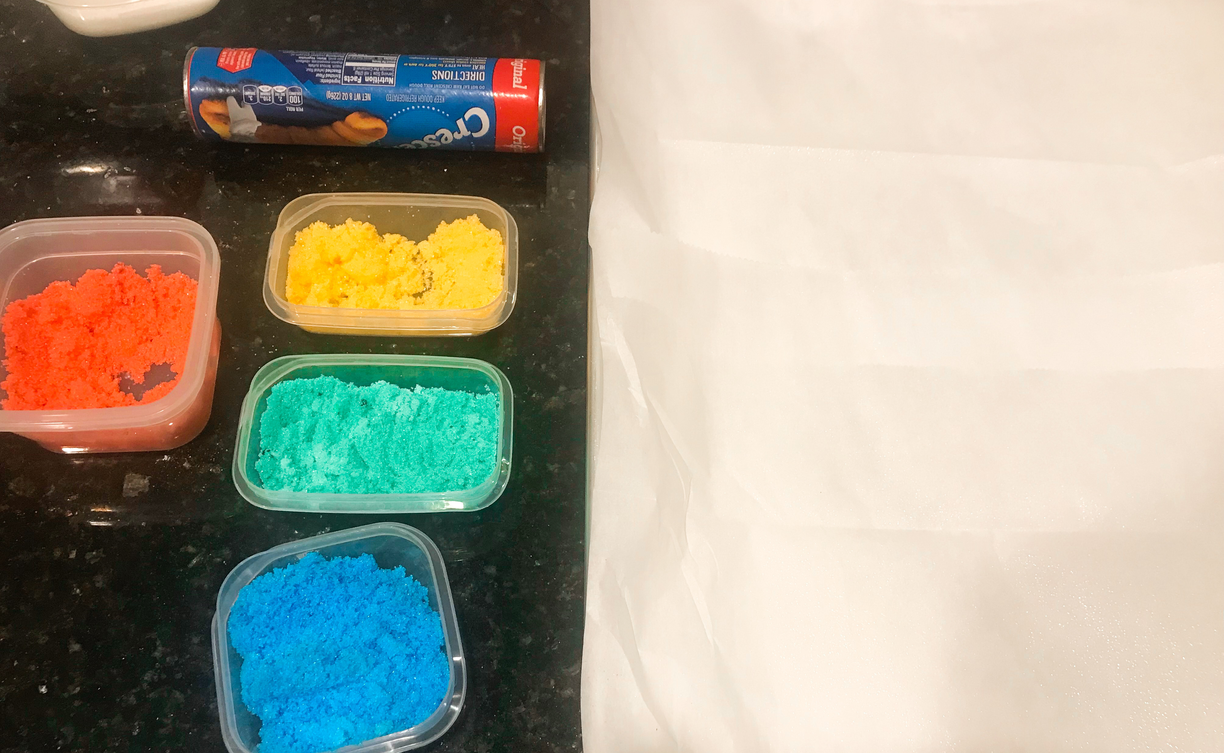 A roll of Pillsbury Crescents, red, green, yellow, and blue sugar in containers and parchment paper on a countertop. 