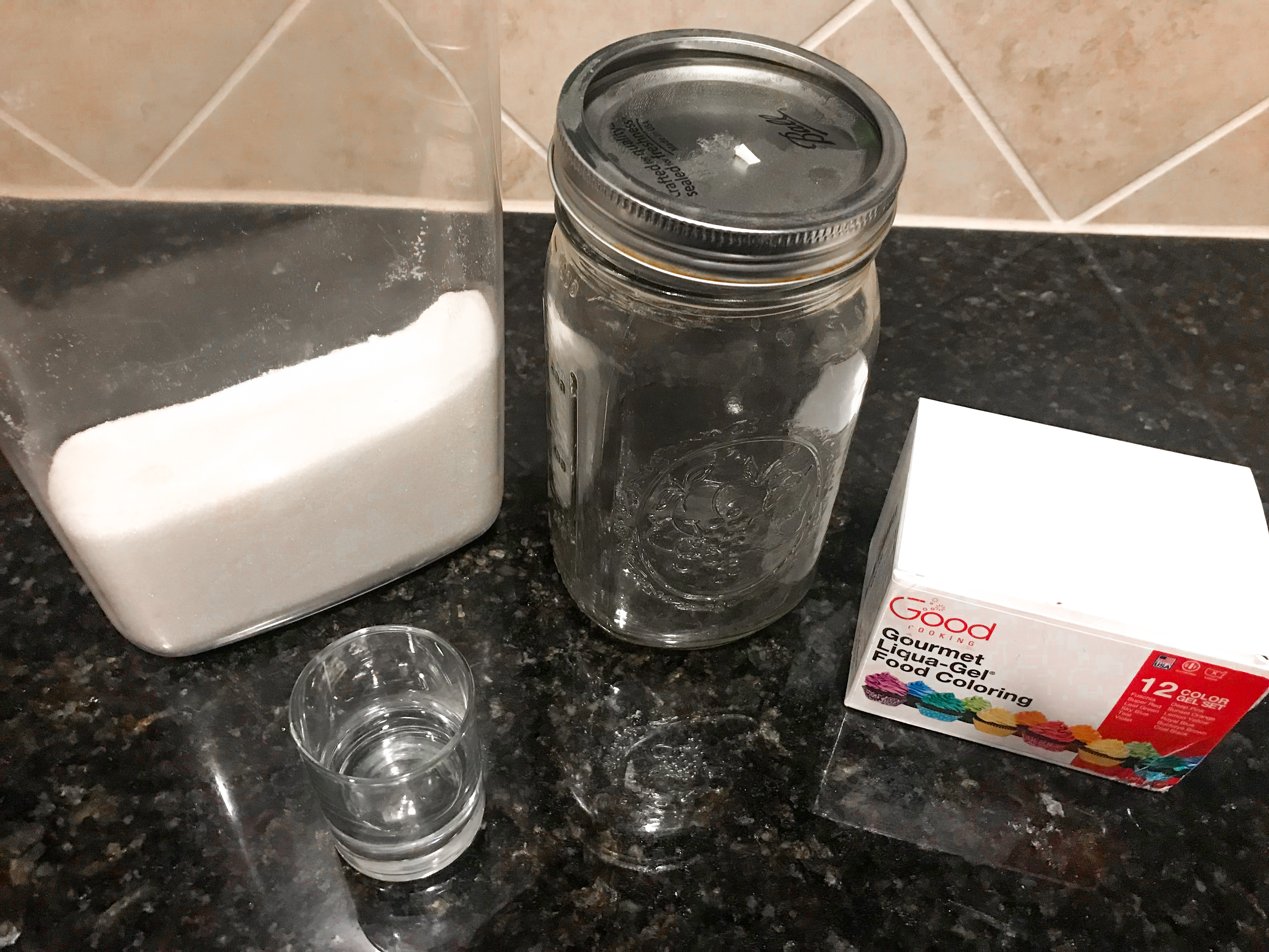 A container full of sugar, a shot glass with a small amount of vodka, an empty mason jar, and a box of food coloring on a countertop. 