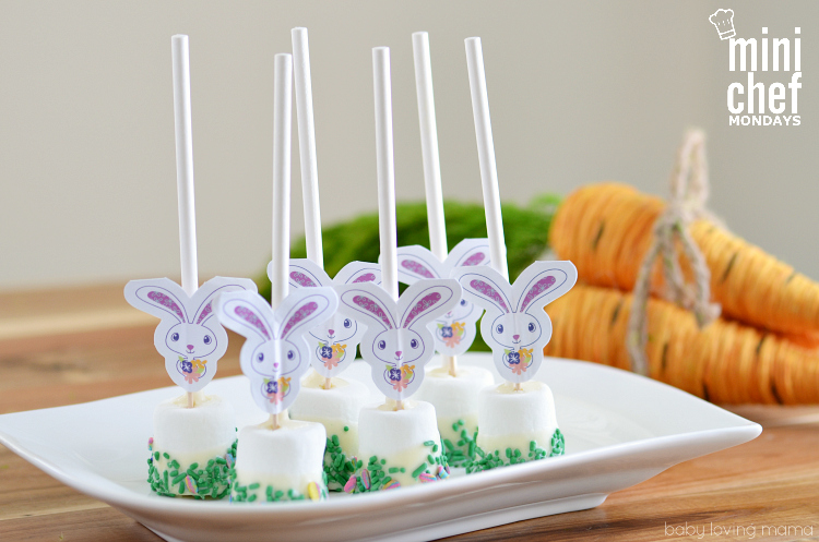 Bunny marshmallow pops dipped in green sprinkles on a white platter with carrot decor in the background. 