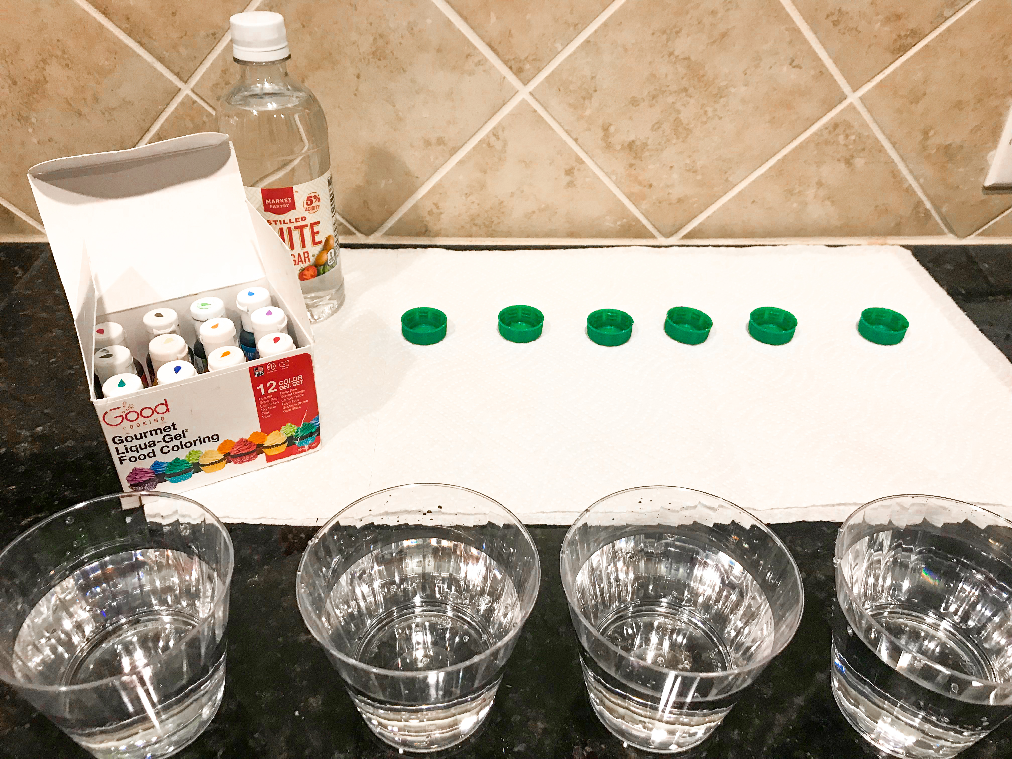 Glasses of water, bottle caps, food coloring, and vinegar on a paper towel. 