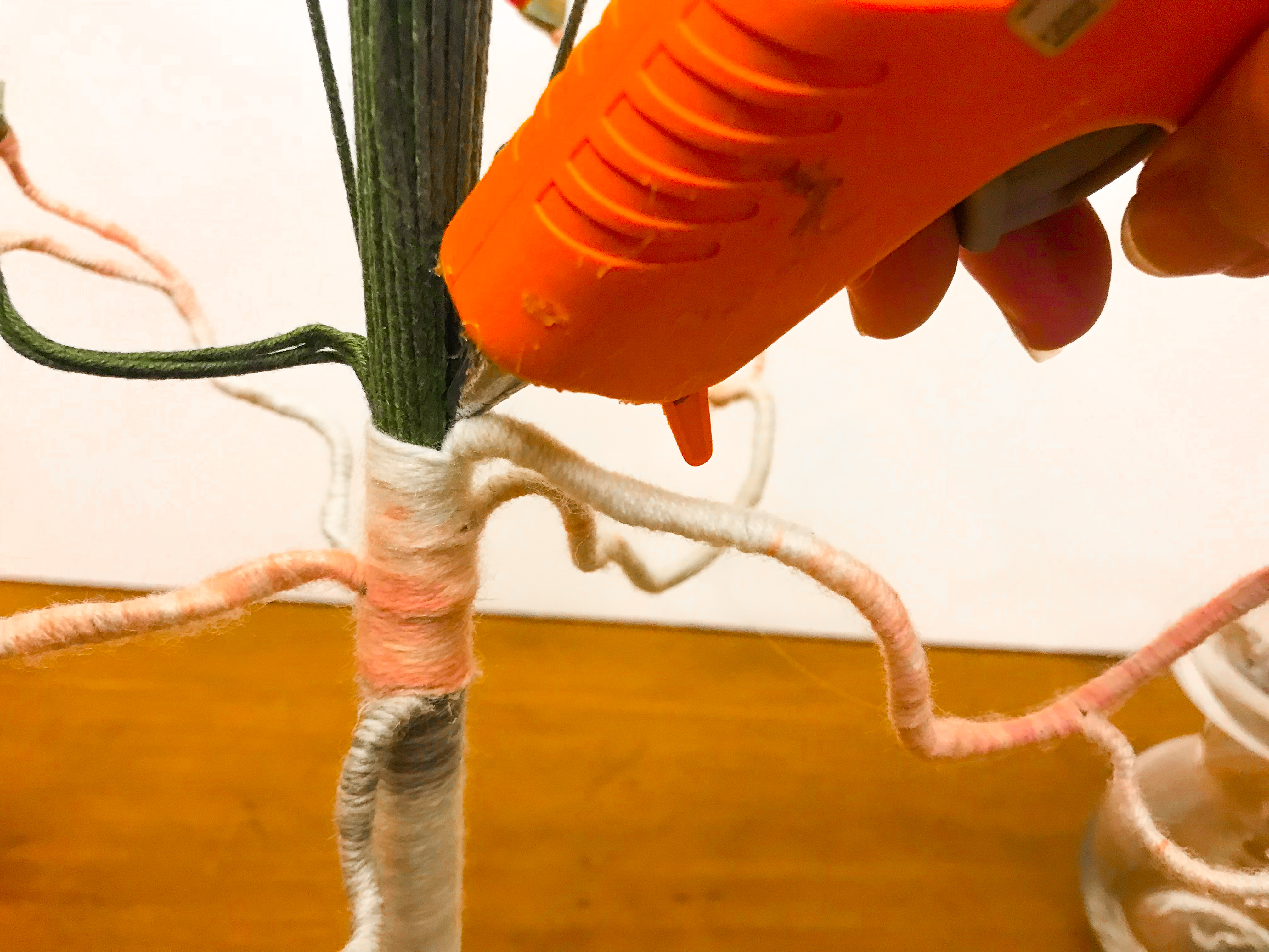 Hot glue gun adding hot glue to junction where yarn wrapped branch meets unwrapped floral stem trunk. 
